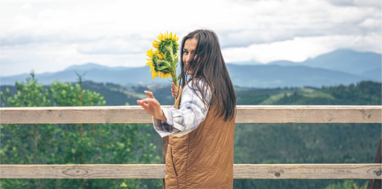 Junge Frau hält Sonnenblumen in der Hand und steht mit dem Rücken zur Kamera und schaut über ihre Schulter. Im HIntergrund ist Landschaft zu erkennen.